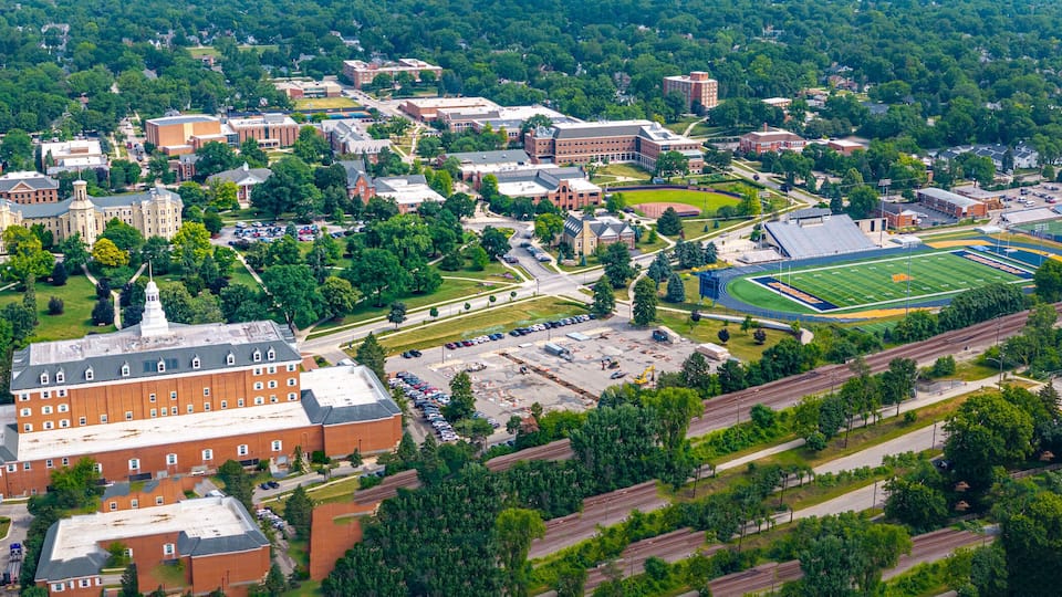 An aerial view of a university campus with multiple buildings, a football field, and surrounding greenery.