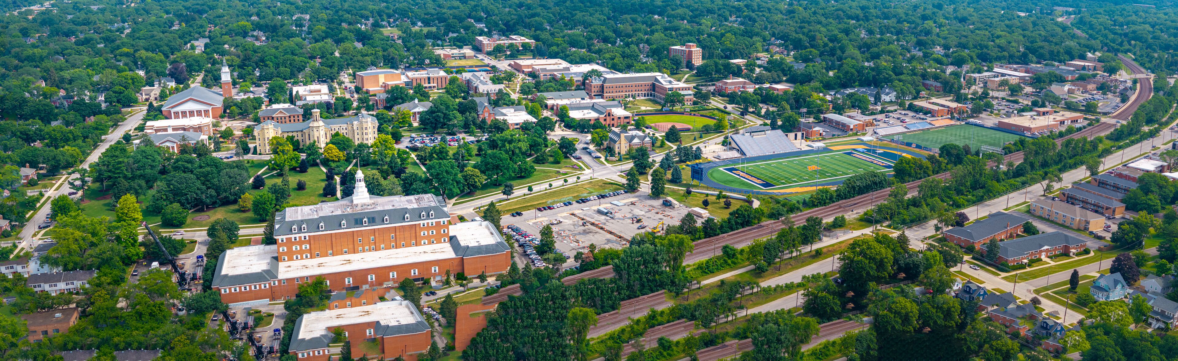 An aerial view of a university campus with multiple buildings, a football field, and surrounding greenery.