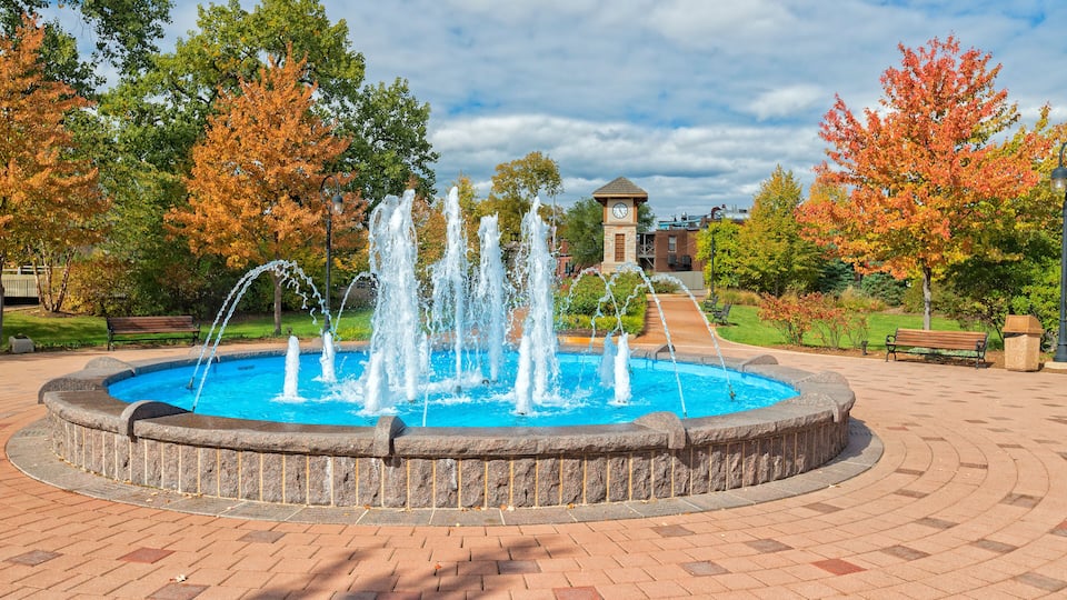 A circular fountain with water jets, surrounded by a brick walkway, with trees and a clock tower in the background.