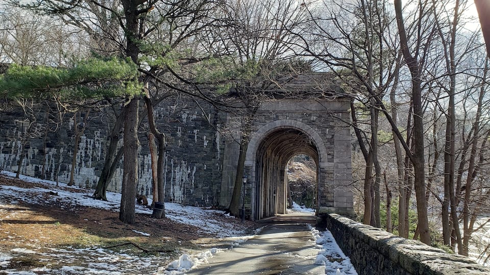 A stone arch bridge in a wooded area with snow on the ground.