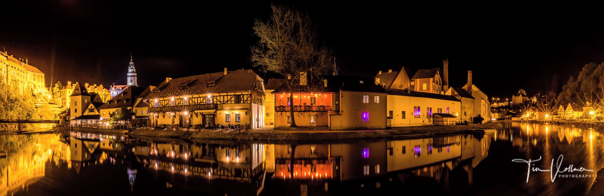 Vista nocturna de un pueblo a orillas del río, con edificios iluminados reflejándose en el agua.