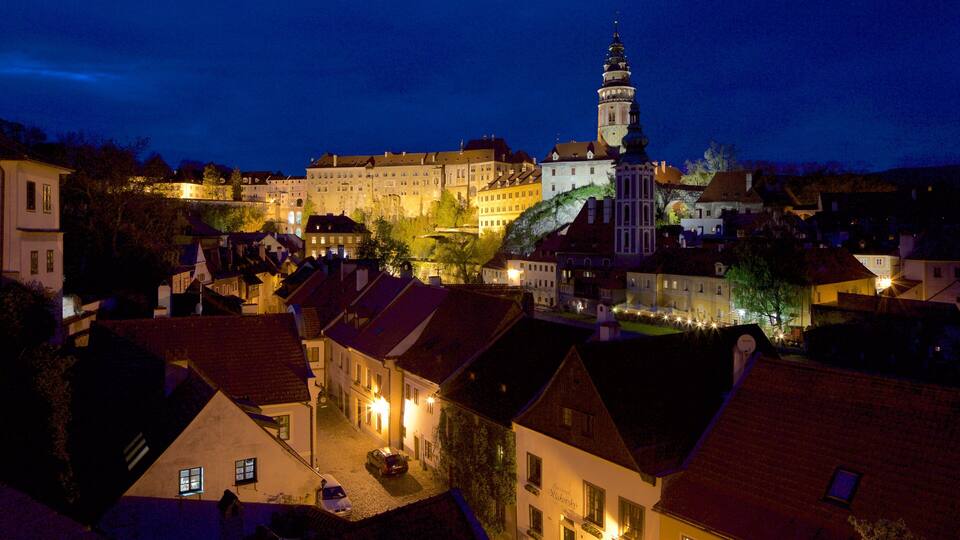 A historic town at night with illuminated buildings, a prominent church tower, and a castle in the background.