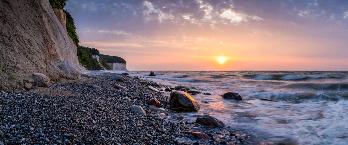 A rocky beach with a cliff, ocean waves, and a sunset.