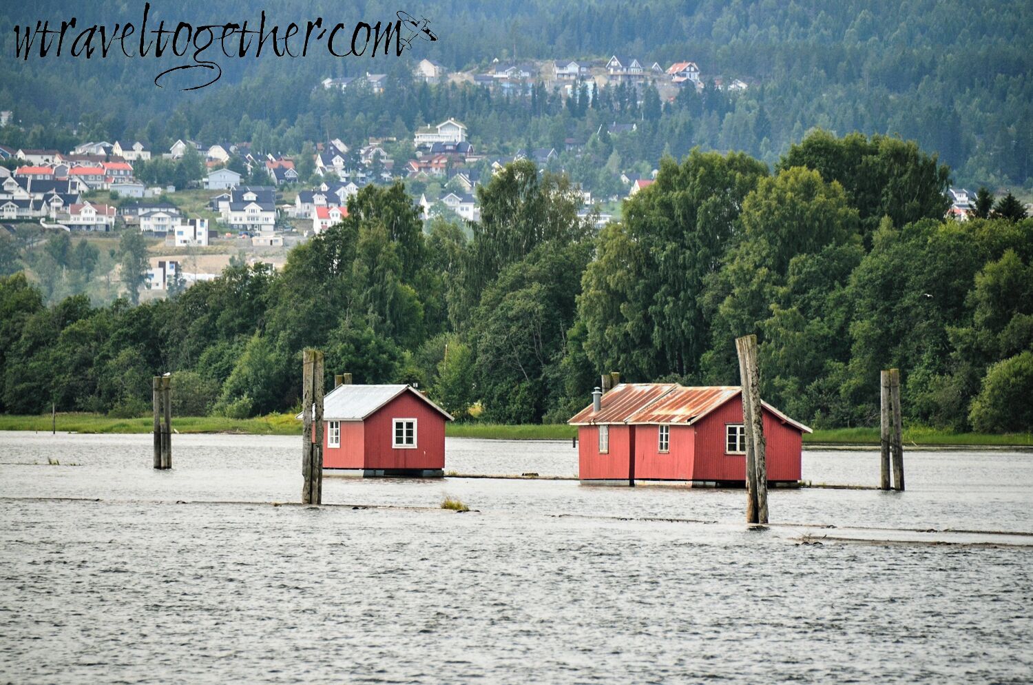 Two red houses on stilts in a flooded area with a forested hill in the background.