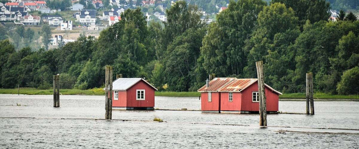 Two red houses on stilts in a flooded area with a forested hill in the background.