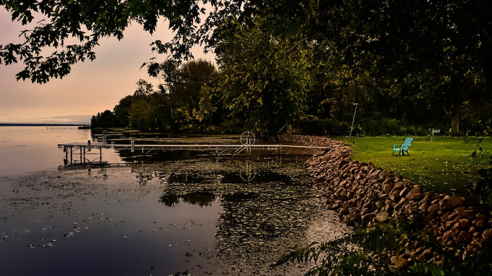A lakeside scene with a dock, a stone wall, and a grassy area with a chair.