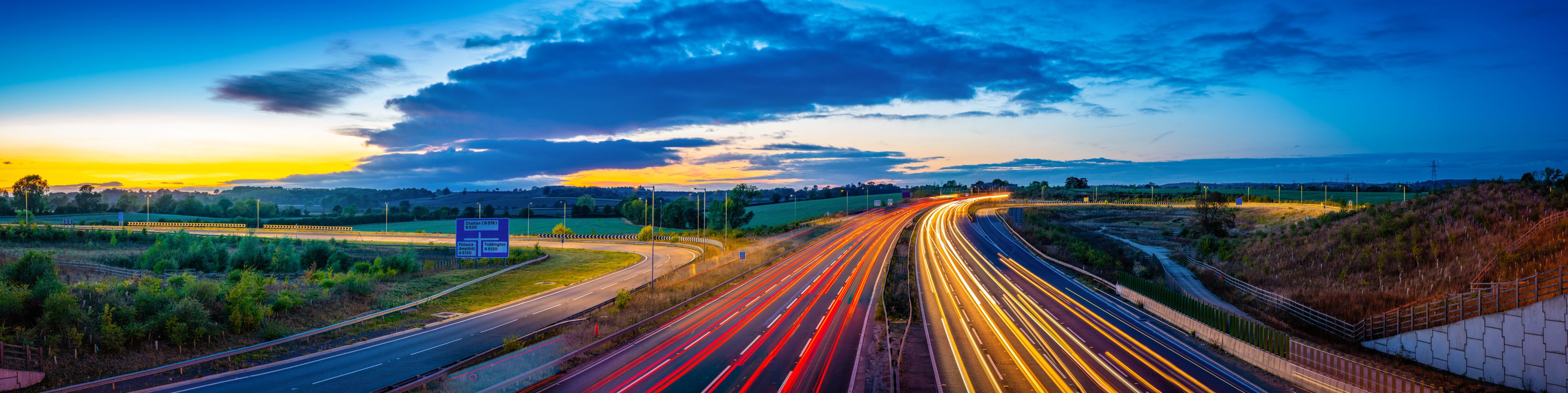 A multi-lane highway with light trails, surrounded by greenery and hills, under a sunset sky.