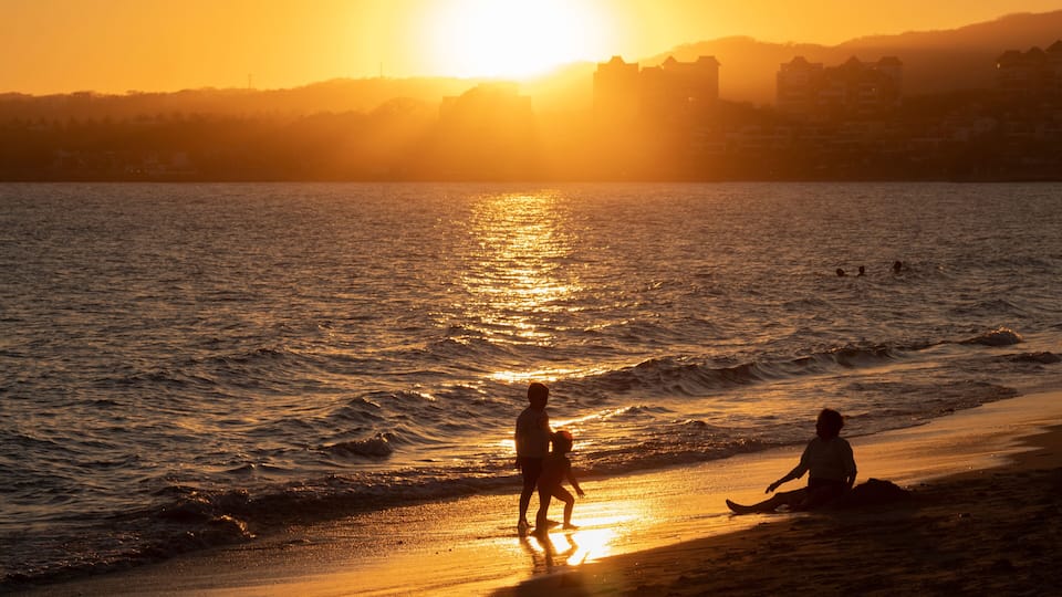 A sunset over a beach with waves, a child playing in the water, and a person sitting on the sand.
