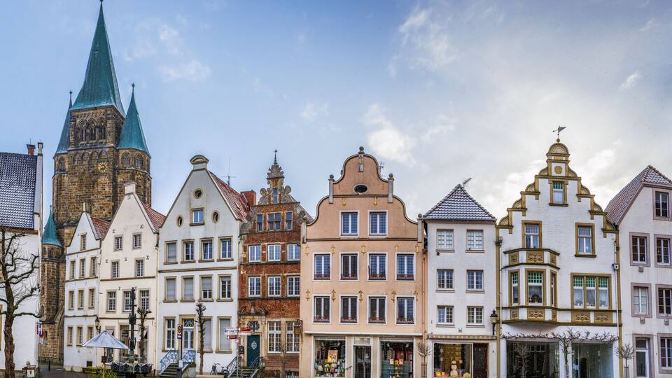 A row of historic European buildings with varied architectural styles, including a church with a green spire.