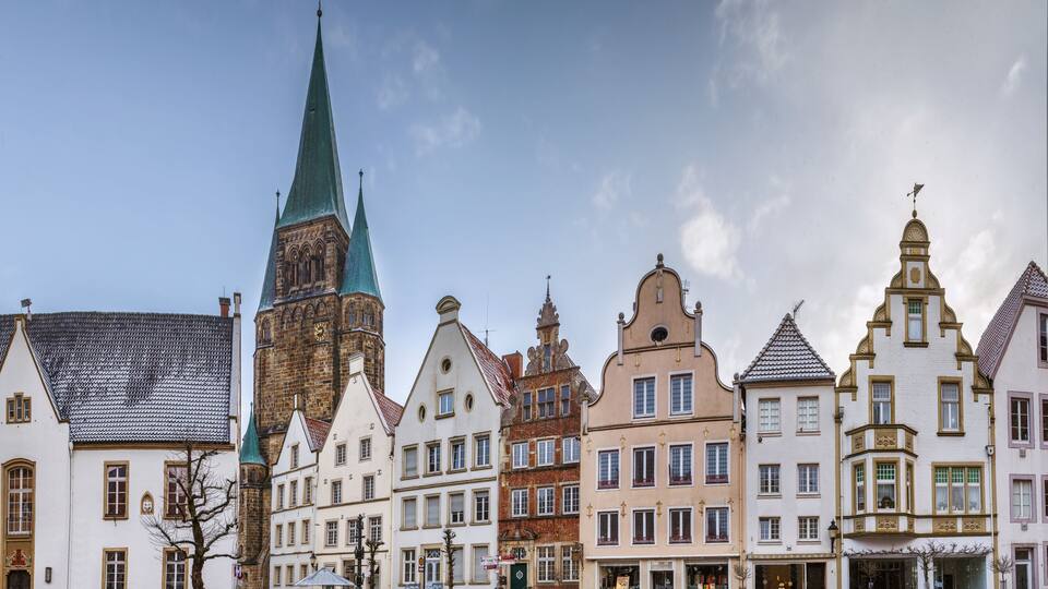 A row of historic European buildings with varied architectural styles, including a prominent church with a green spire.