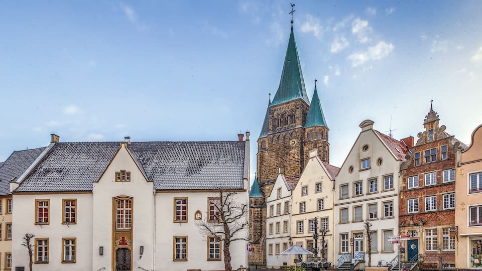 A historic European town square with traditional buildings, a prominent church tower, and a clear blue sky.