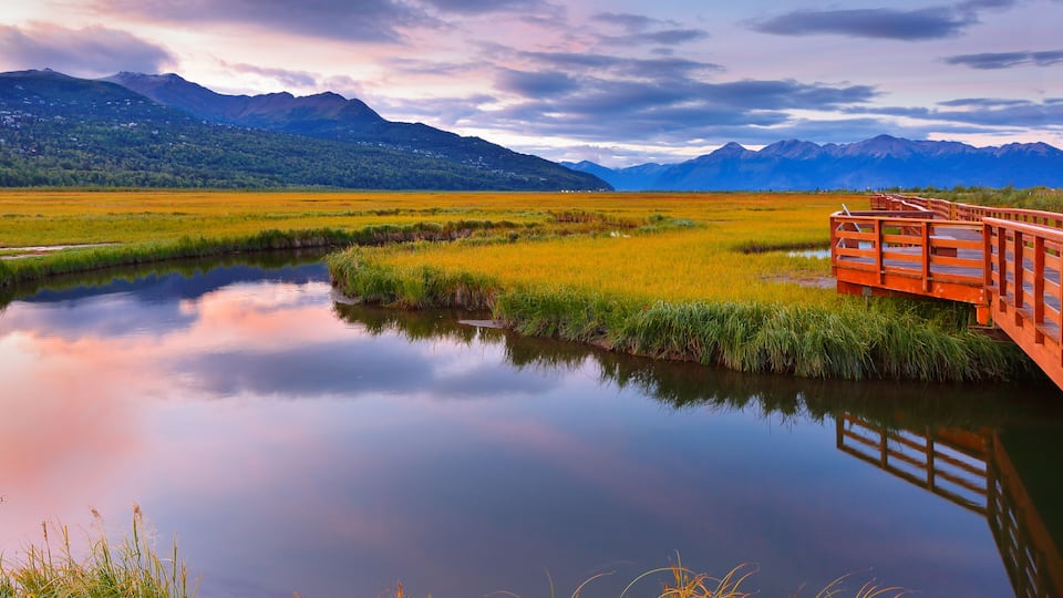 A calm lake with a wooden bridge, surrounded by green fields and mountains under a cloudy sky.