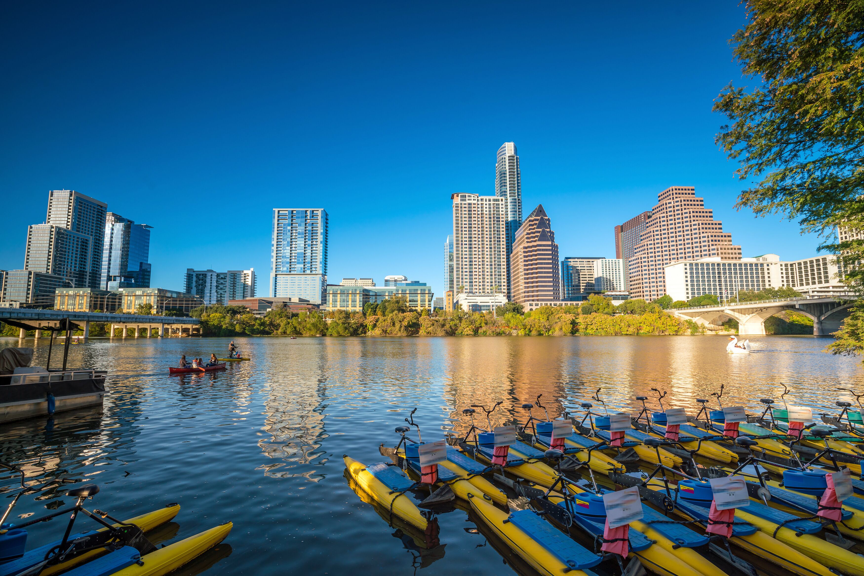 A river with yellow and blue kayaks lined up on the shore, a bridge, and a city skyline with tall buildings in the background.
