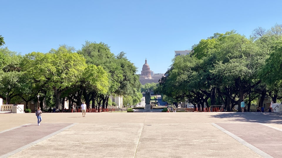 A wide open plaza with a large statue in the center, surrounded by trees and buildings in the background.