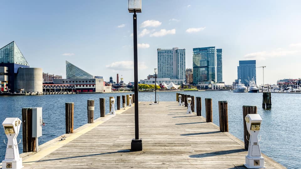 A wooden pier extending into a body of water with modern buildings in the background.