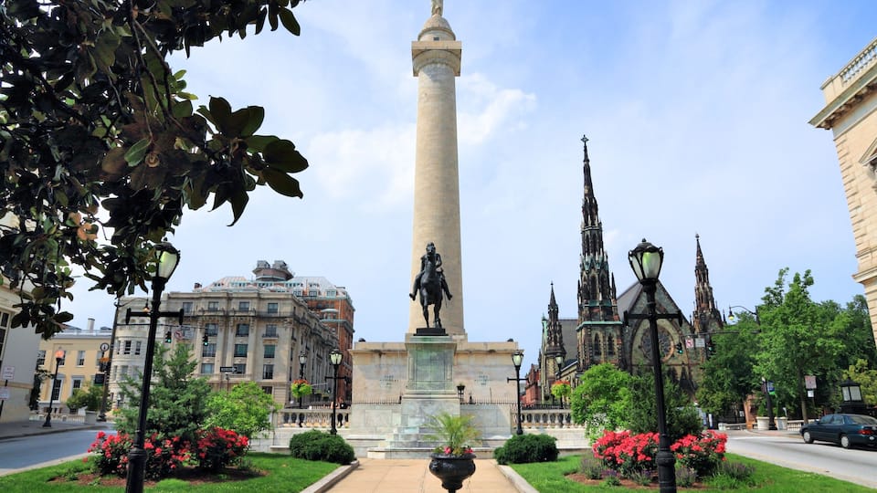 A monument with a statue, surrounded by well-maintained gardens and historic buildings.