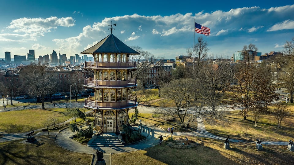 A tower with a green roof, surrounded by a park with trees and a city skyline in the background.