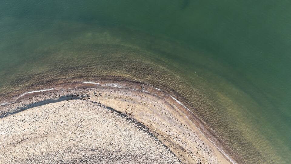 An aerial view of a beach with clear water, a sandy shore, and visible footprints.