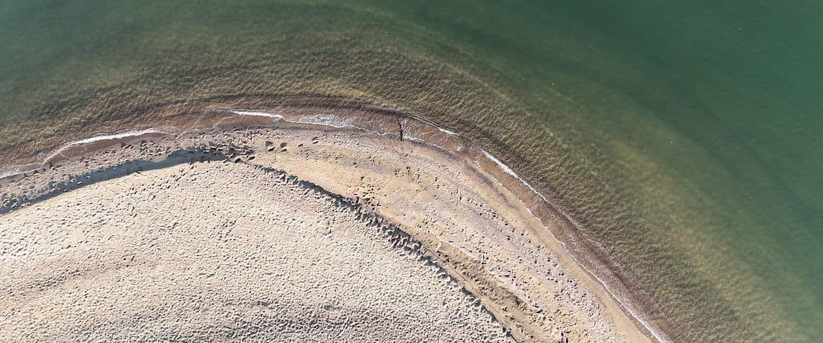 An aerial view of a beach with clear water, a sandy shore, and visible footprints.