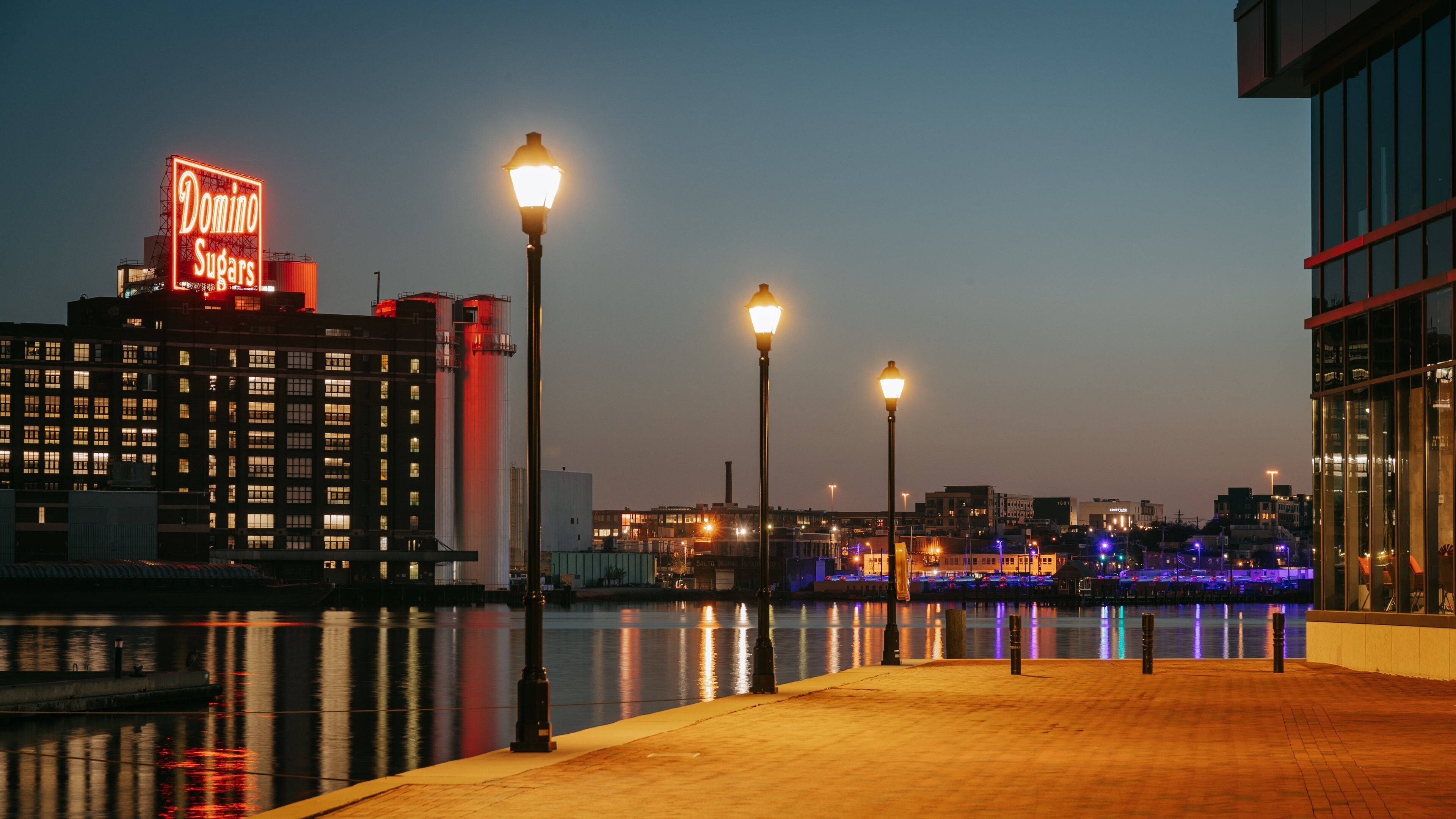 A waterfront promenade at dusk with illuminated street lamps and a prominent Domino Sugars sign reflecting on the water.
