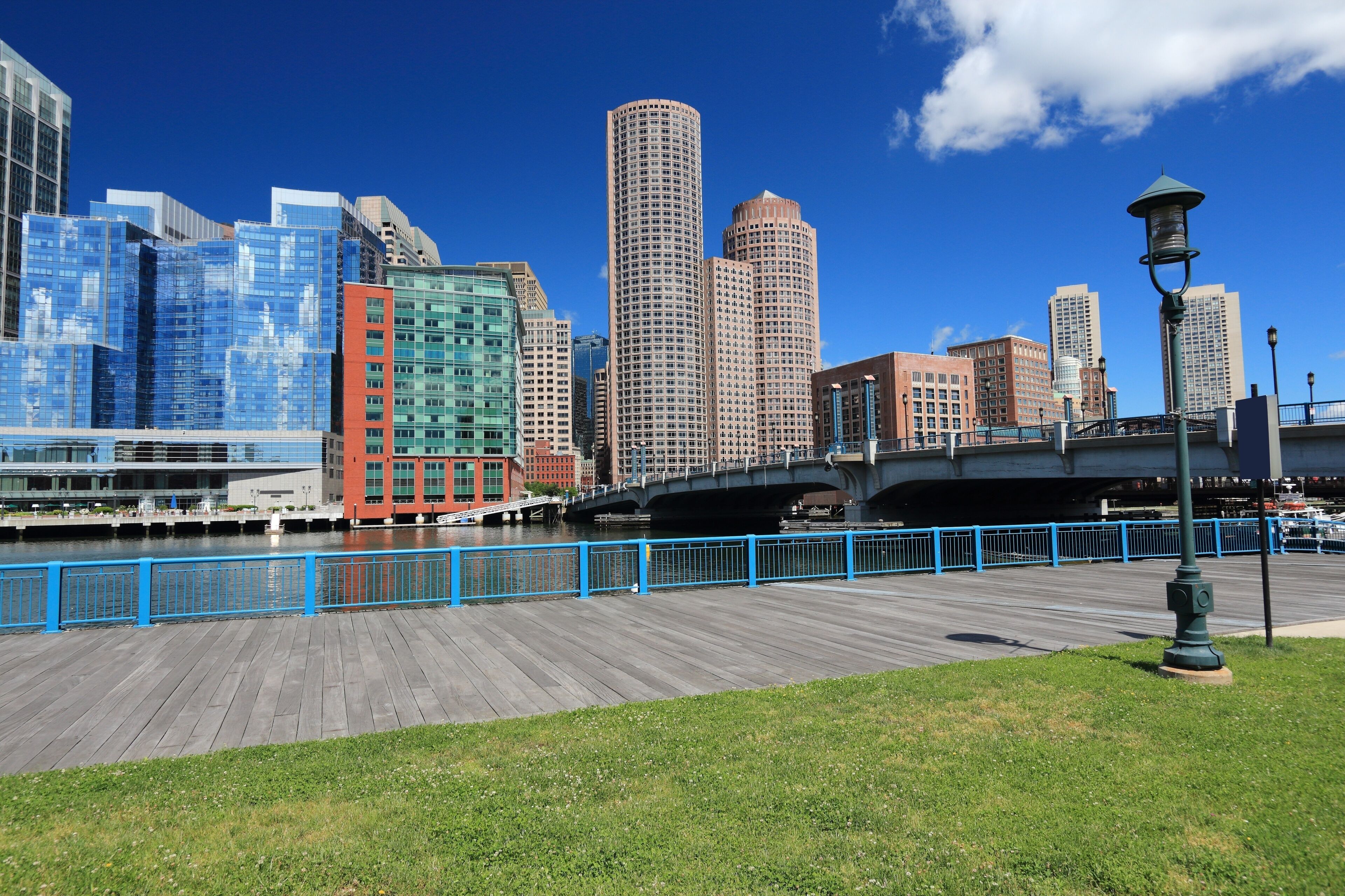 A waterfront promenade with a wooden boardwalk, a bridge, and a city skyline with modern buildings.