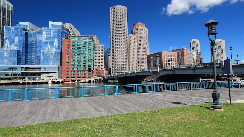 A waterfront promenade with a wooden boardwalk, a bridge, and a city skyline with modern buildings.