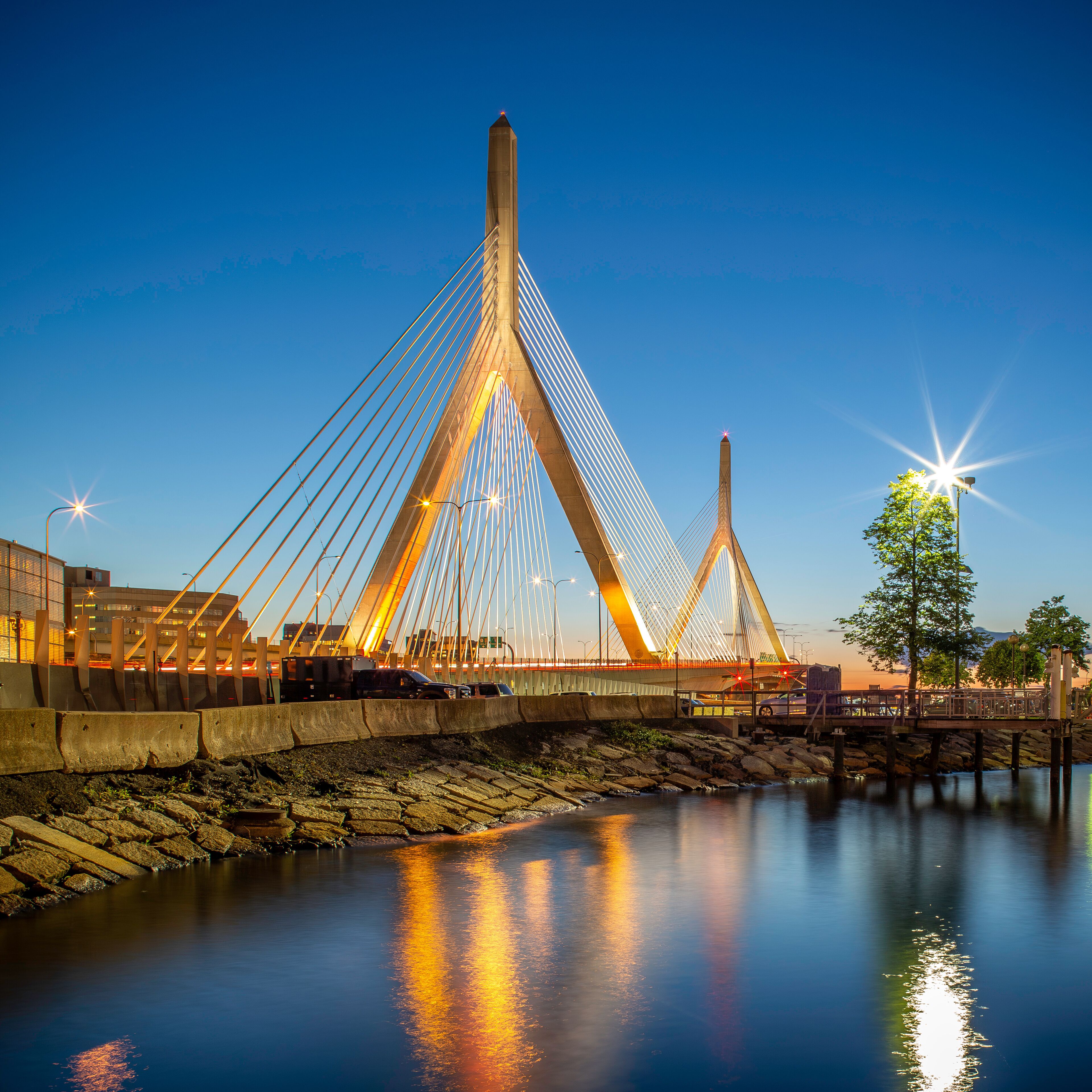 A cable-stayed bridge with a central tower and cables extending to the sides, over a calm river with a rocky shore.