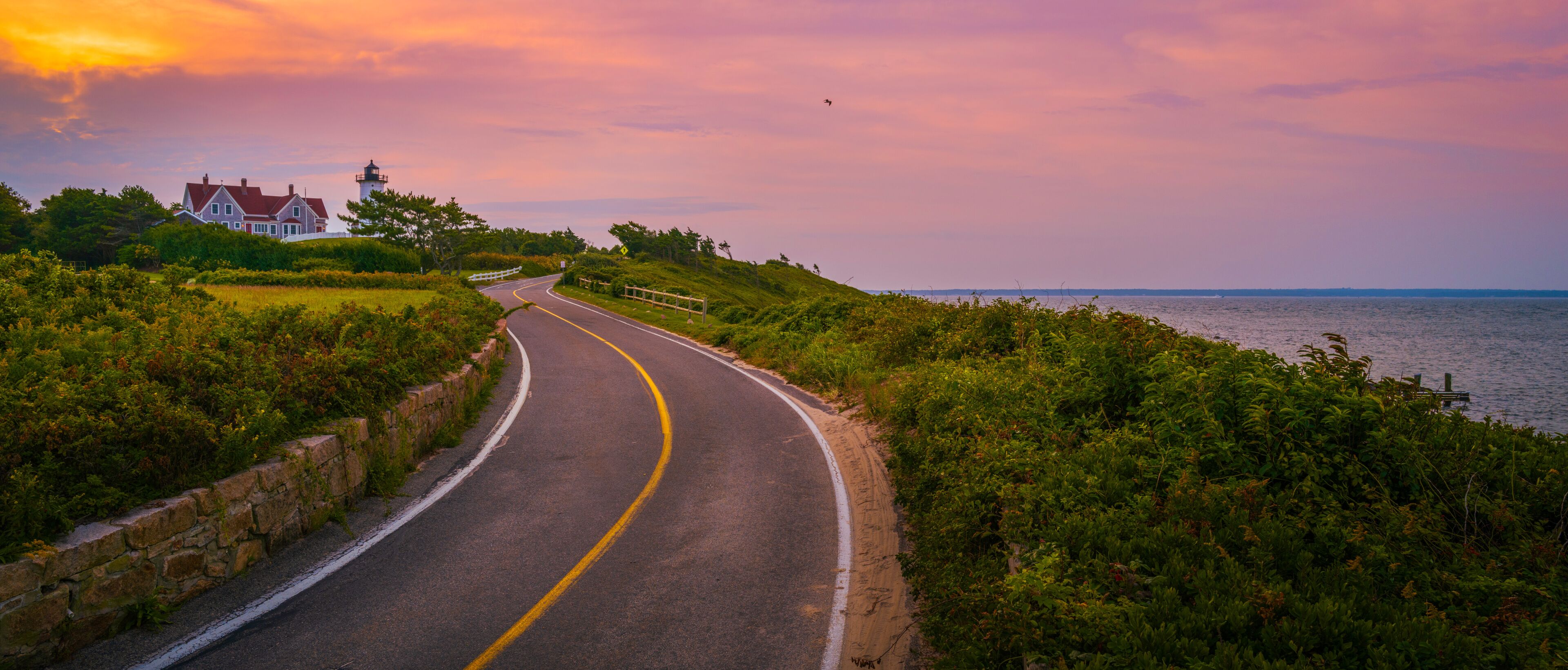 A coastal road curving along a grassy embankment with a lighthouse and a house in the distance.