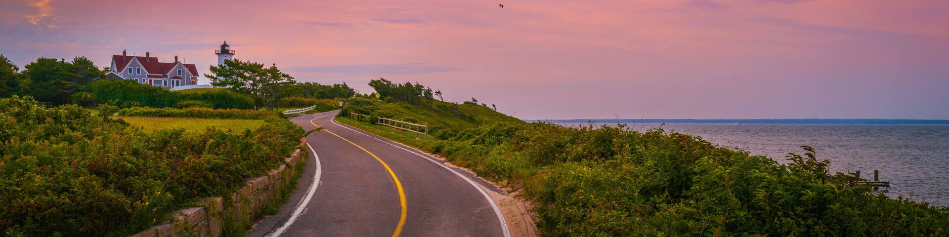 A coastal road curving along a grassy embankment with a lighthouse and a house in the distance.