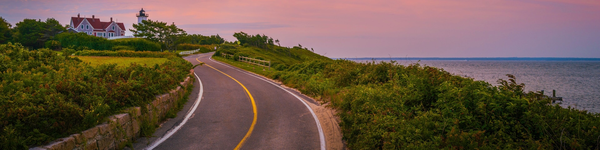 A coastal road curving along a grassy embankment with a lighthouse and a house in the distance.