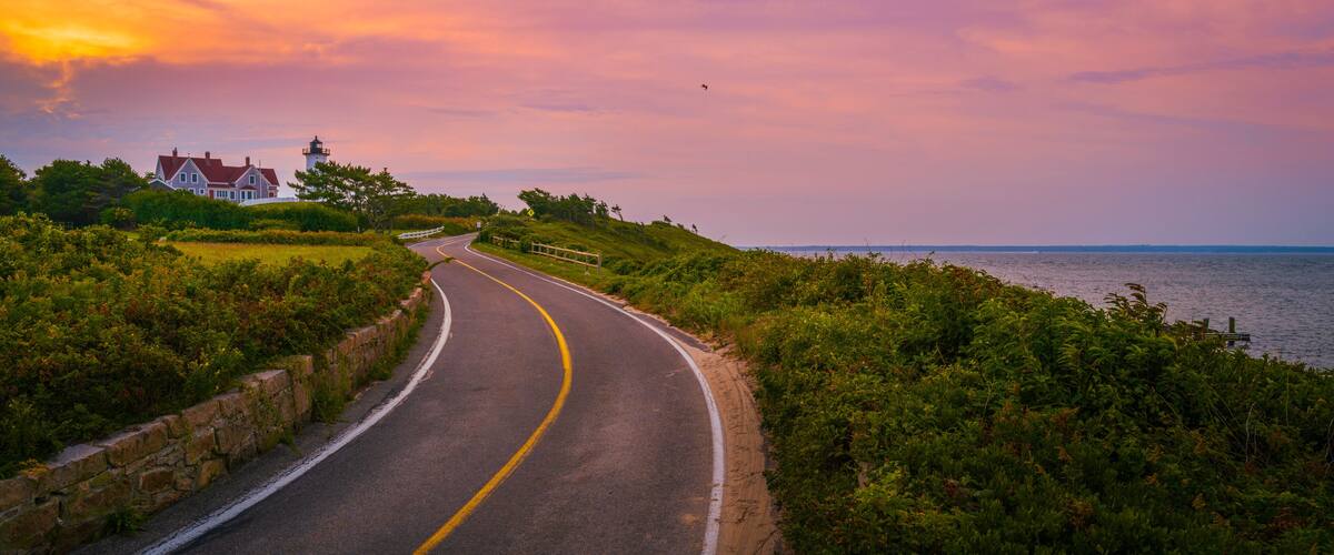 A coastal road curving along a grassy embankment with a lighthouse and a house in the distance.
