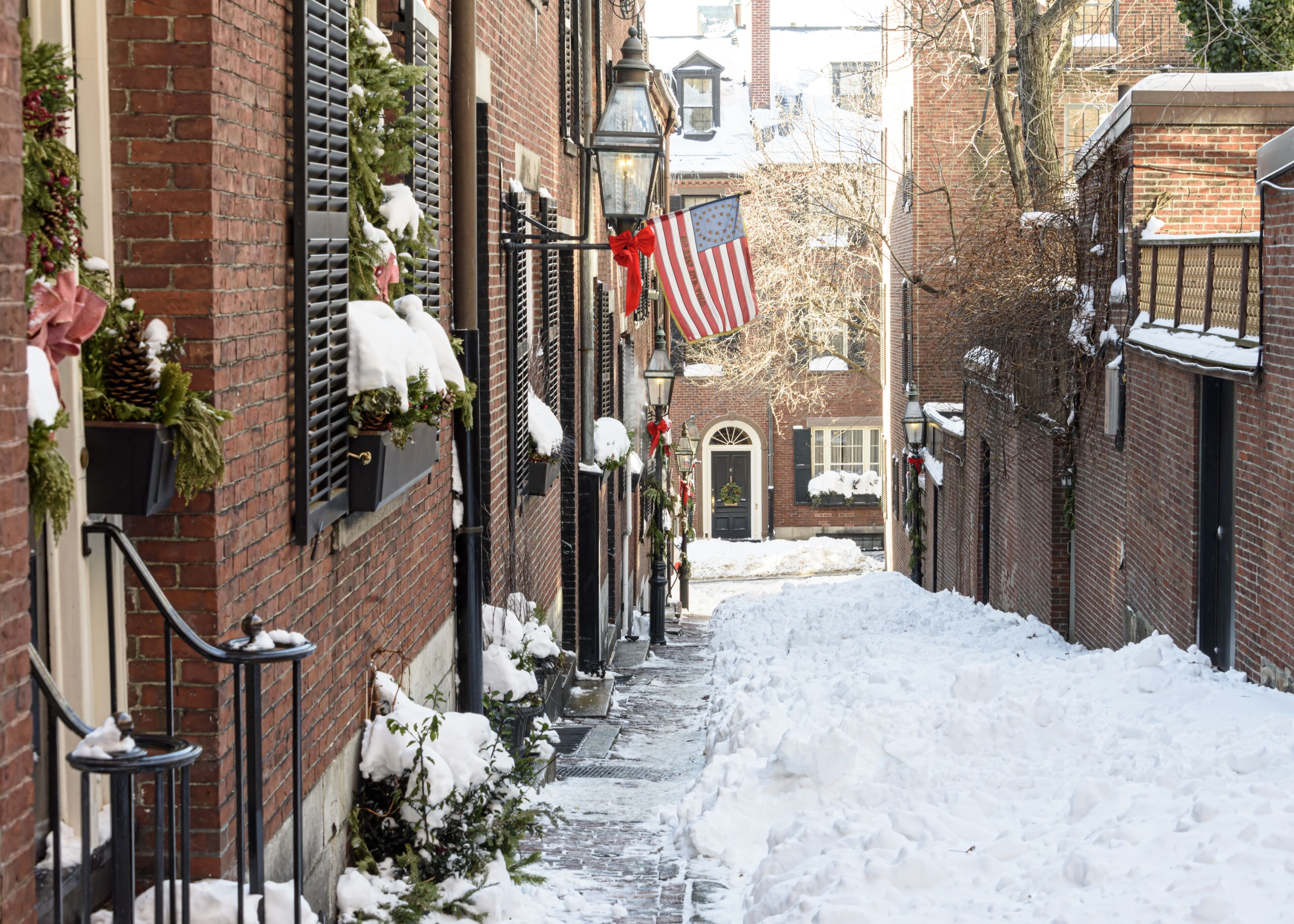 A snow-covered alley with brick buildings, American flags, and Christmas decorations.