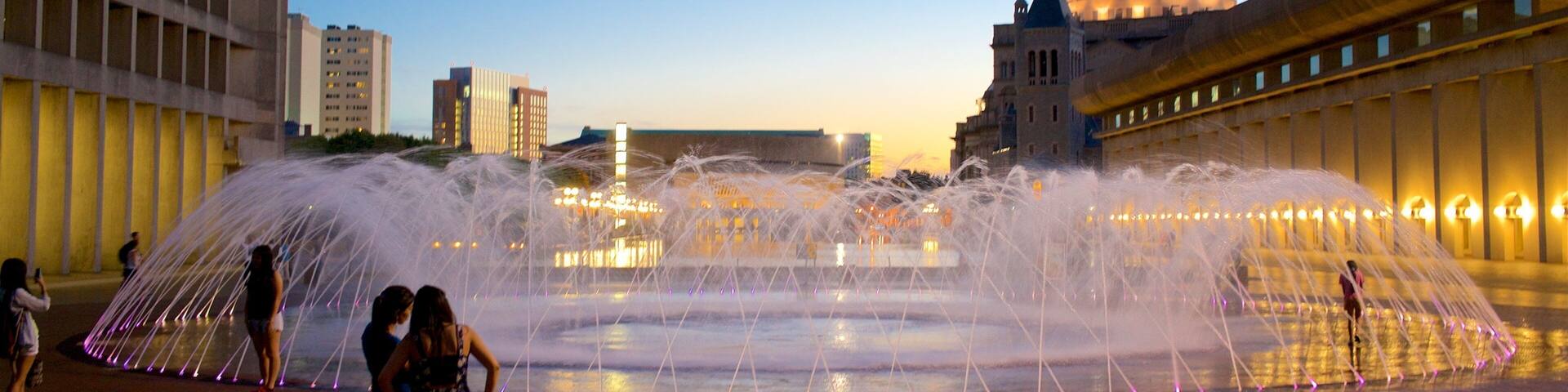 A large water fountain with multiple jets, a domed building in the background, and modern buildings around.