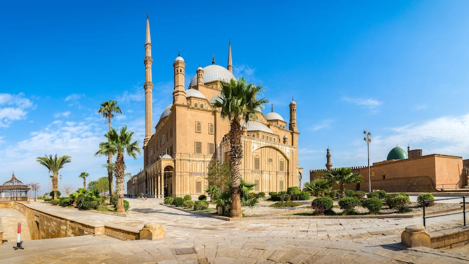 A historic mosque with multiple domes and minarets, surrounded by palm trees and a paved courtyard.