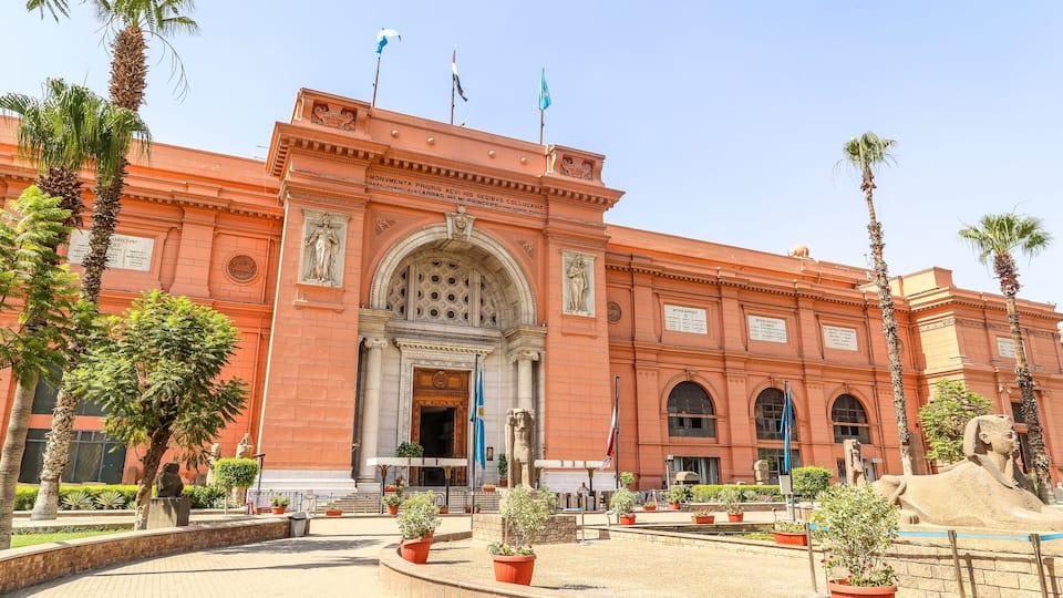 A large red building with a central archway, palm trees, and statues.