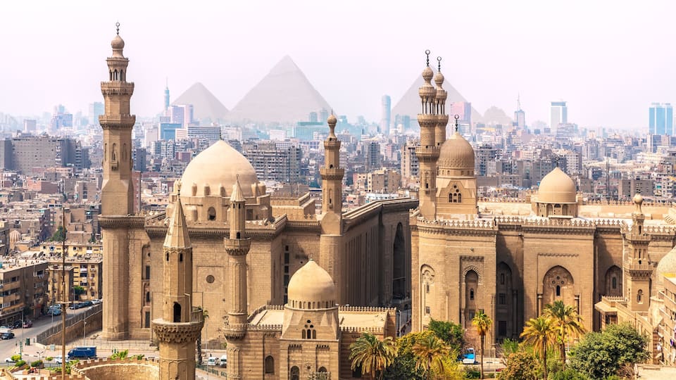A historic mosque with multiple domes and minarets, set against a backdrop of the pyramids and a modern cityscape.