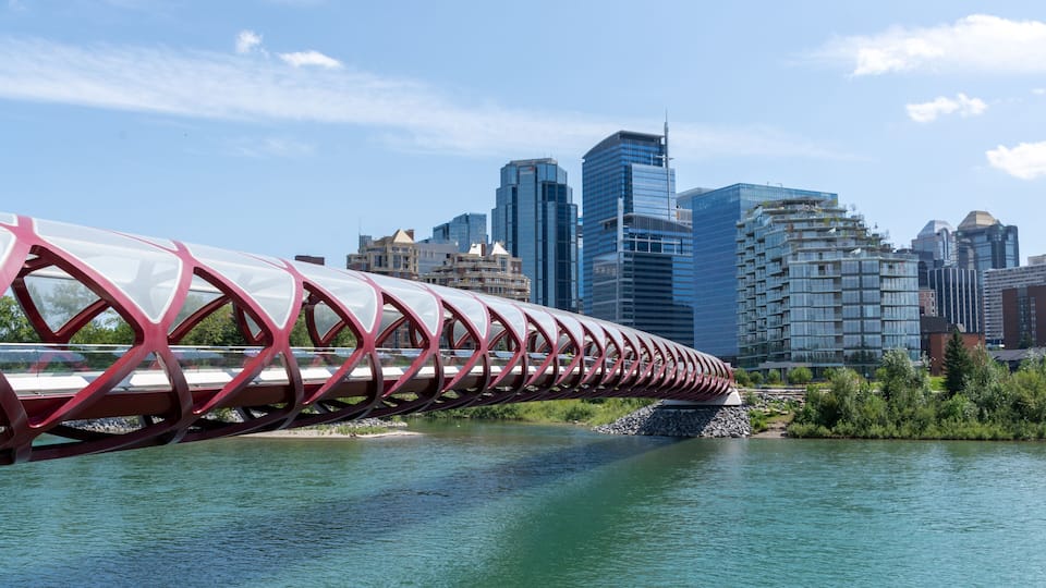 A modern bridge with a red and white design over a river, with a city skyline in the background.