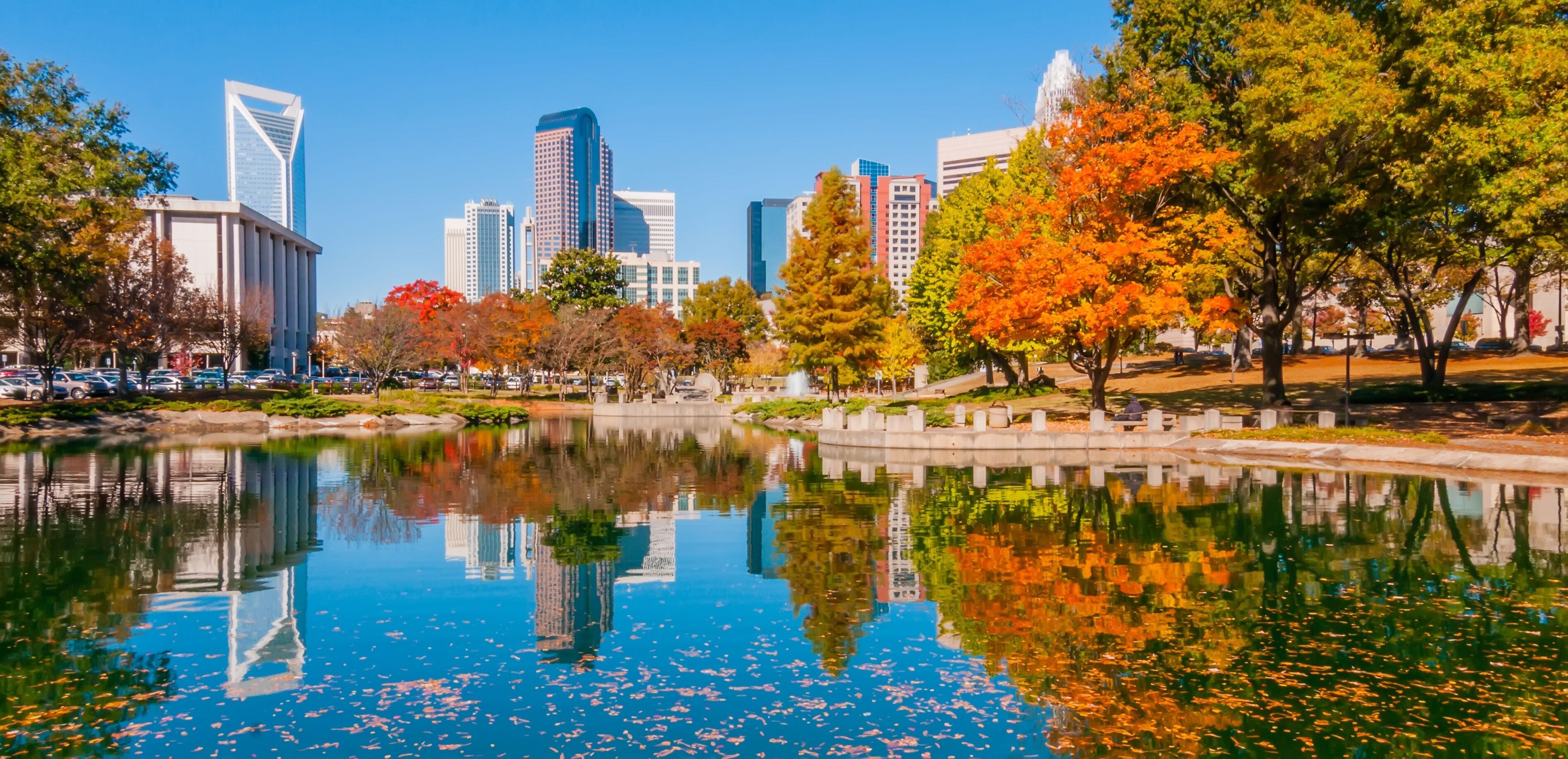 A cityscape with autumn trees, a reflective lake, and modern skyscrapers.
