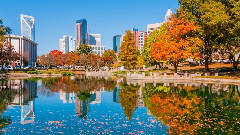 A cityscape with autumn trees, a reflective lake, and modern skyscrapers.