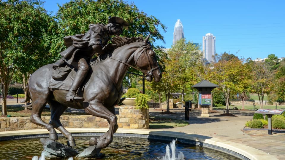 A bronze equestrian statue in a park with a fountain, trees, and a distant cityscape.