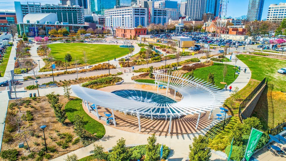 A modern urban park with a unique white pavilion, green lawns, and surrounding city buildings.