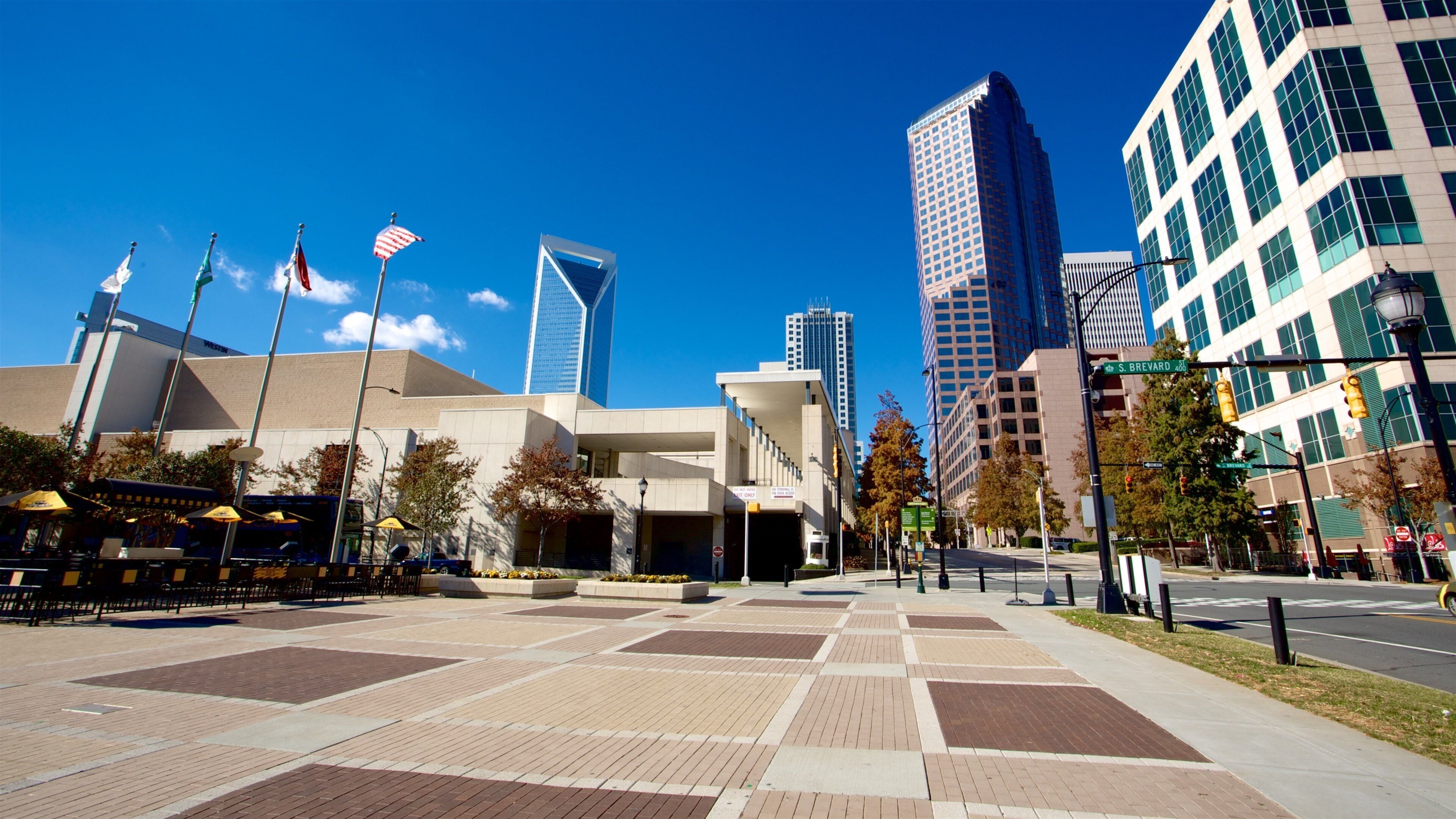 A city square with modern buildings, flags, and a clear blue sky.