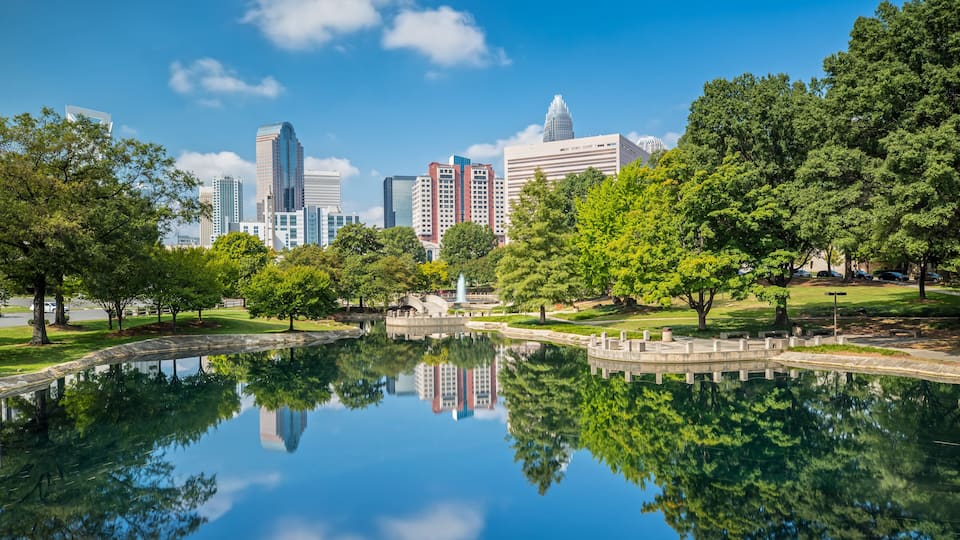 A city skyline reflected in a calm lake, surrounded by lush green trees and a park with a fountain.