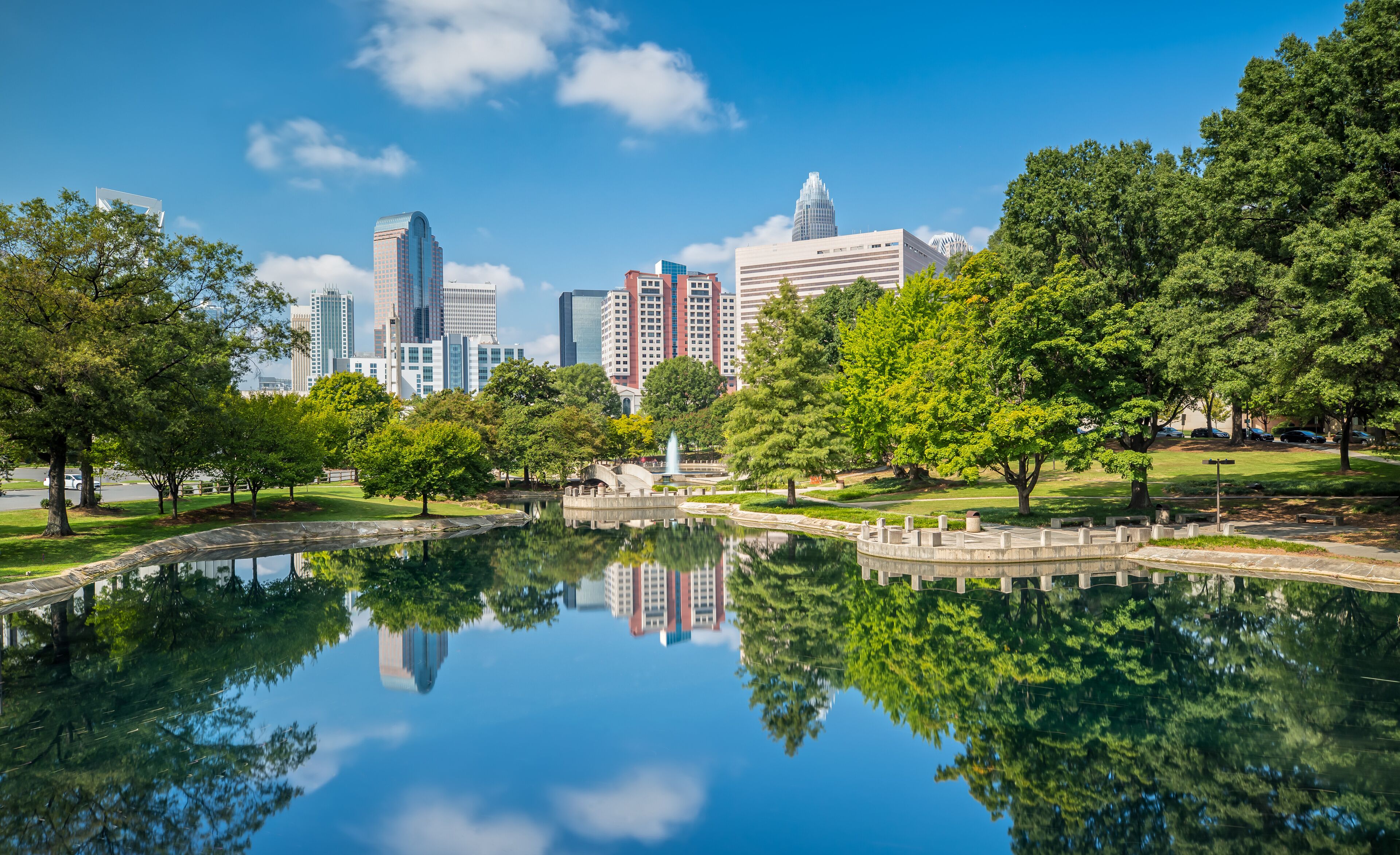 A city skyline reflected in a calm lake, surrounded by lush green trees and a park with a fountain.