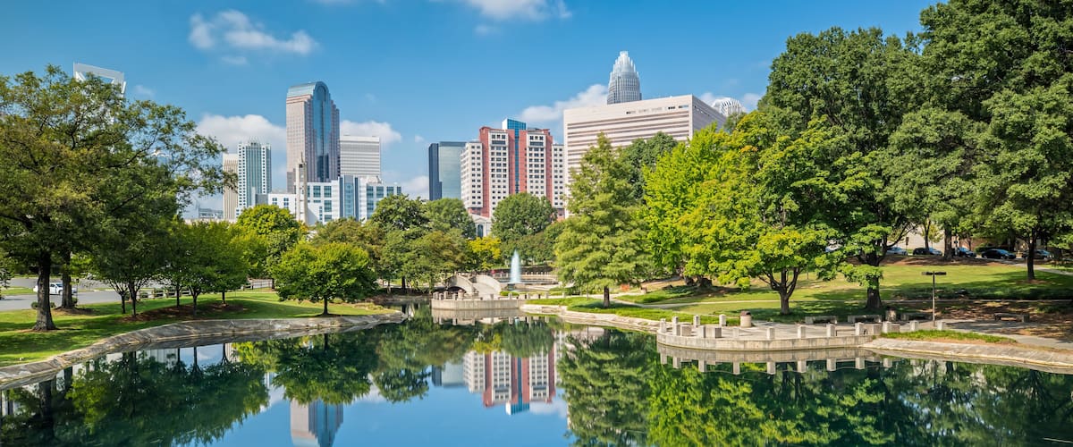 A city skyline reflected in a calm lake, surrounded by lush green trees and a park with a fountain.