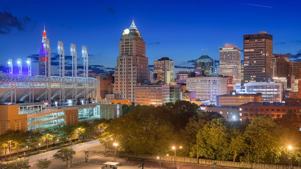 A city skyline at dusk with illuminated buildings, a prominent tower, and a stadium with a retractable roof.