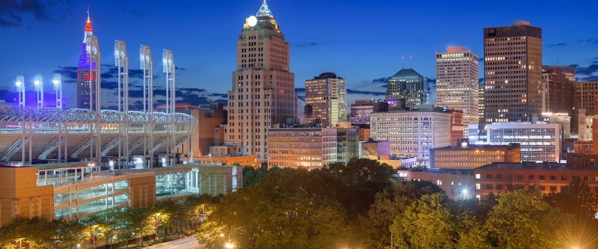 A city skyline at dusk with illuminated buildings, a prominent tower, and a stadium with a retractable roof.