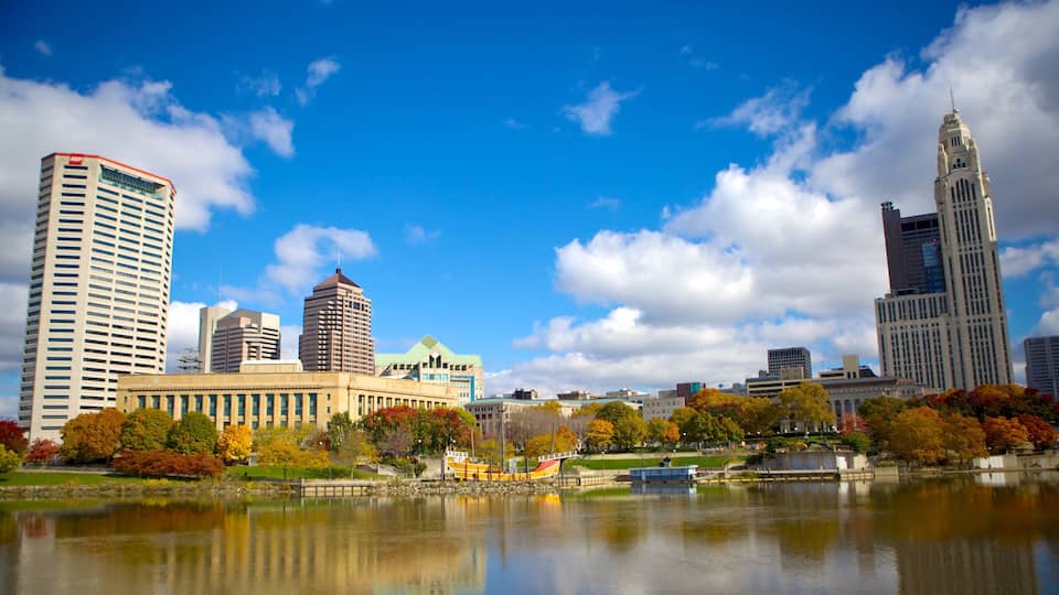 A city skyline with tall buildings, a lake, and autumn-colored trees.
