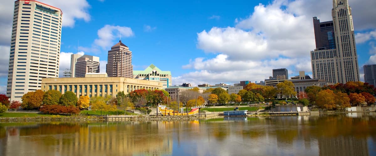 A city skyline with tall buildings, a lake, and autumn-colored trees.