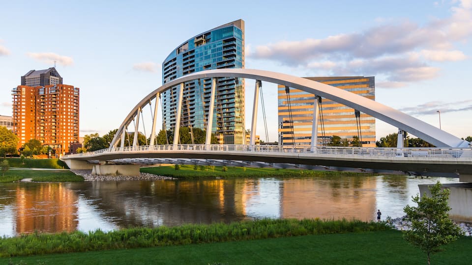A modern bridge with a unique arch design spanning a river, surrounded by urban buildings and greenery.
